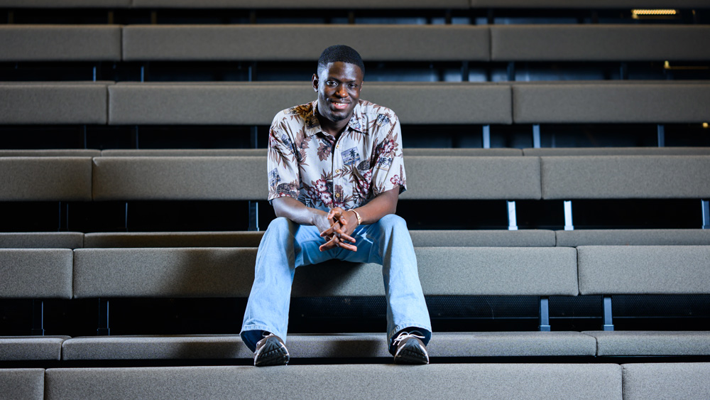 Kwesi Afrifa sits on the bleachers of a theatre with hands clasped. The rows of bleachers create strong horizontal lines.