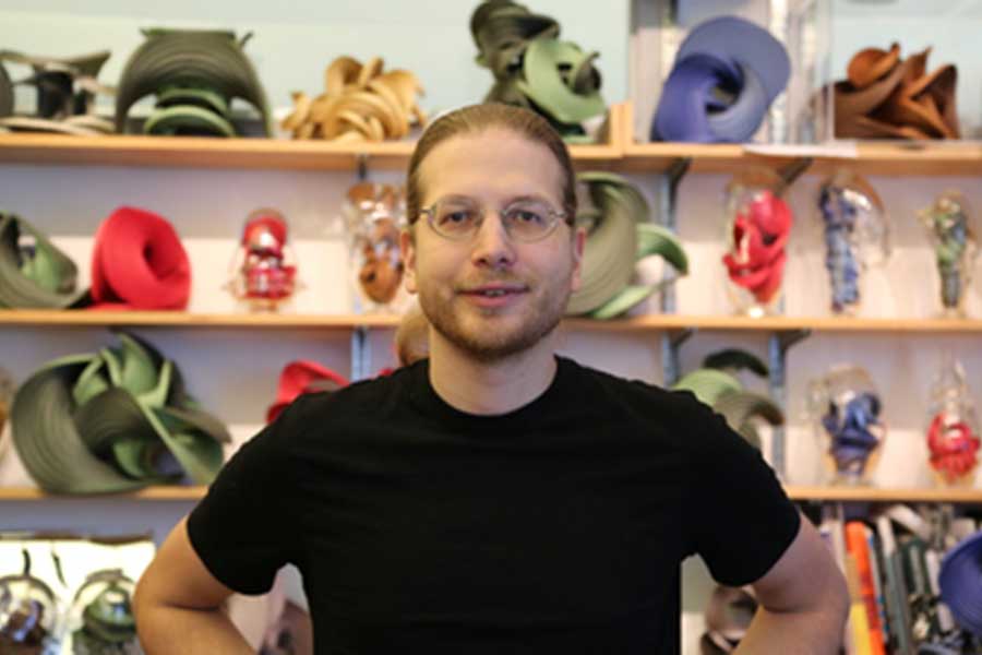 Erik Demaine portrait with unique origami sculptures on shelves in background