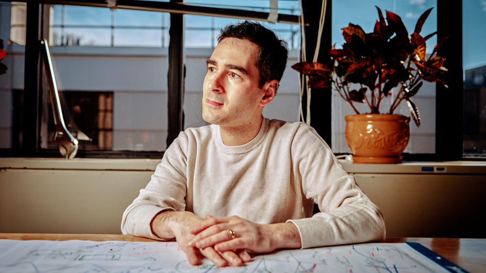 Rafael Jaramillo sits in his office and looks to the side. A large wrench sits on the window sill. The desk is covered in white paper with many drawings and notes on it. 