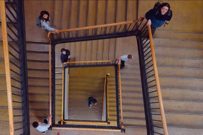 Aerial view of a stairwell spiraling down, as 6 singers look up and sing while walking up the stairs
