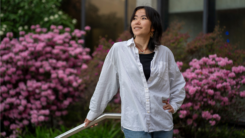 Cindy Xie smiles outside and looks up and to the side. The background has blooming pink rhododendrons.