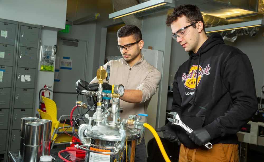 Aly kombargi and Niko Tsakiris are in the lab wearing safety googles and  working with a tabletop device with many tubes and valves.