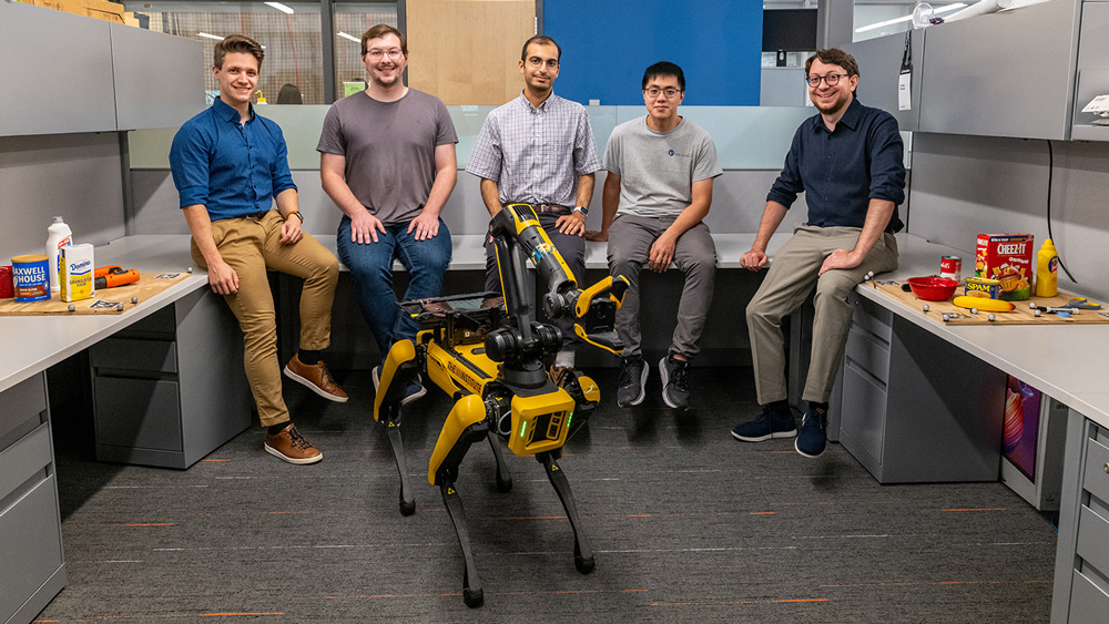 5 guys are in the office around their quadruped robot Spot.