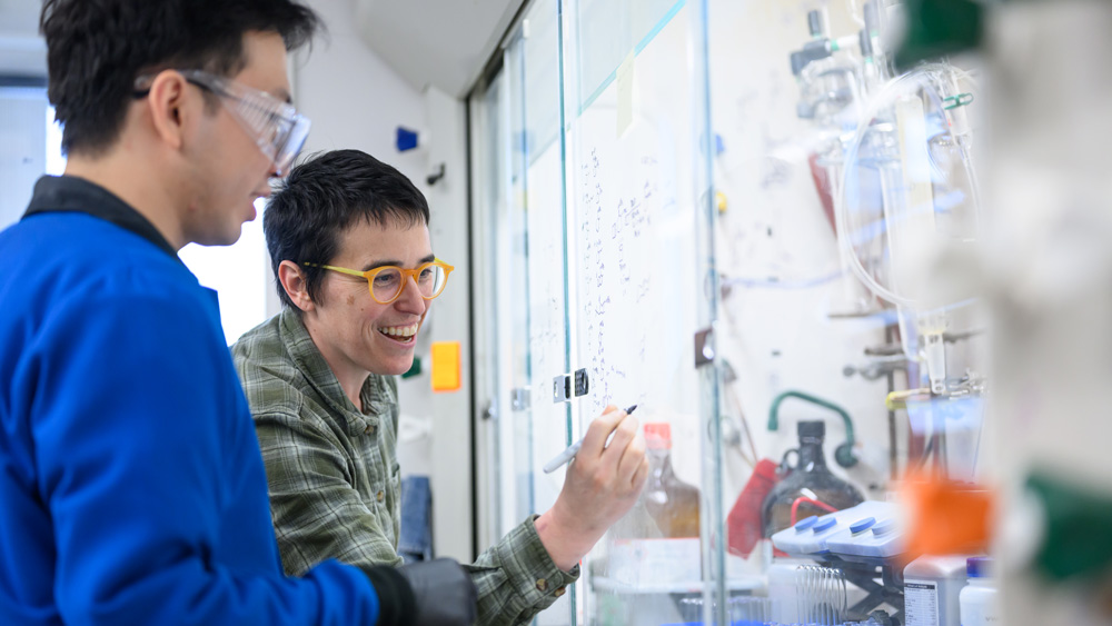 Wendlandt writes on a window in a lab, with student nearby