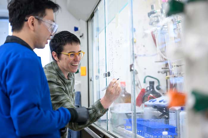 Wendlandt writes on a window in a lab, with student nearby