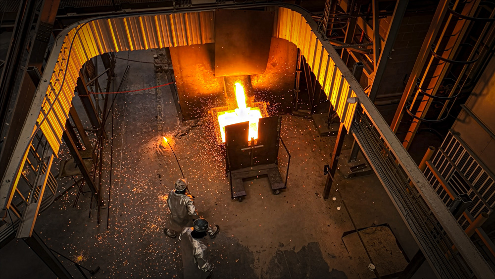 A blast of molten steel causes dazzling sparks as two people in protective suits, one with a long pole, stand back.